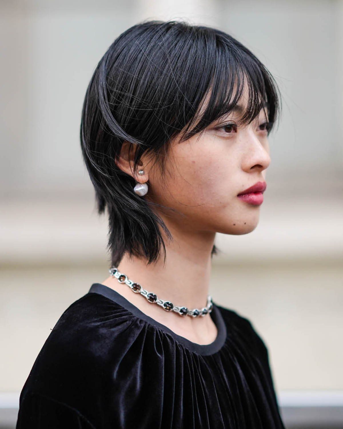 Mulher com cabelo estilo bixie cut, usando brincos grandes e colar prateado, veste blusa preta de veludo.