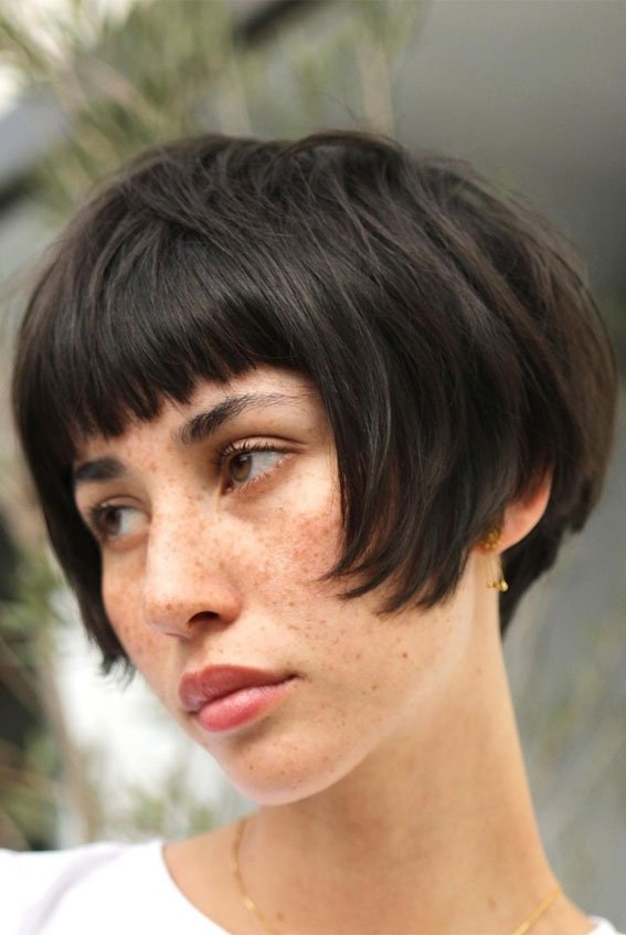 Pessoa com cabelo bixie cut escuro e franja curta, olha para o lado, com expressão tranquila.