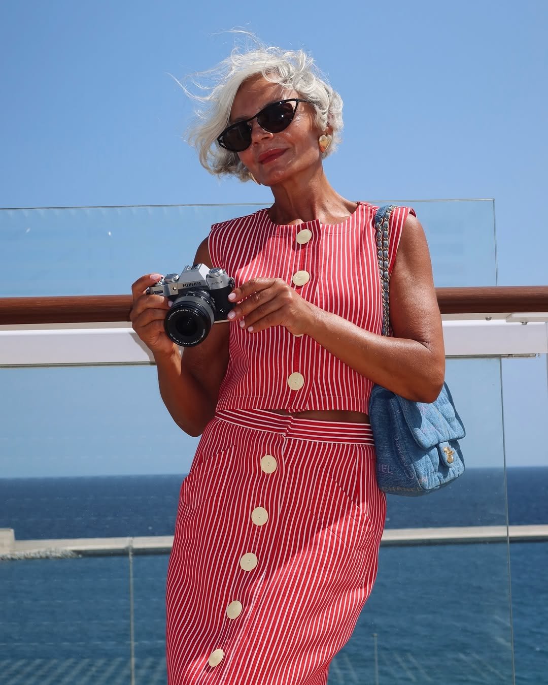 Usuária com vestido listrado vermelho e branco segura uma câmera. Ao ombro, bolsa matelassê azul. Fundo de mar e céu.