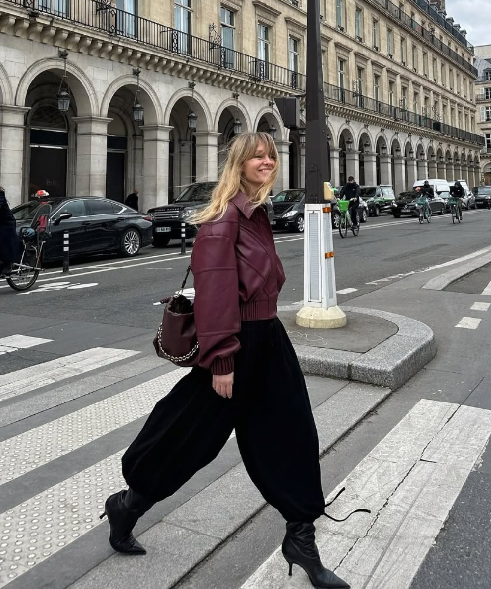 Mulher atravessa rua usando jaqueta vinho, calça larga preta e bota preta feminina, com edifícios ao fundo.