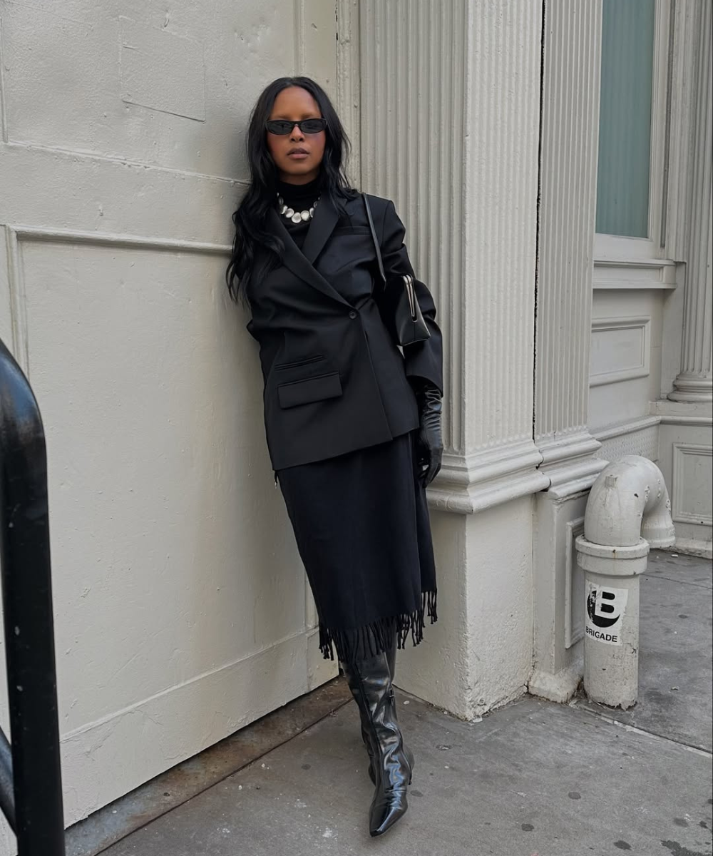 Mulher com óculos escuros e colar de pérolas, usando blazer preto e bota preta feminina em calçada urbana.