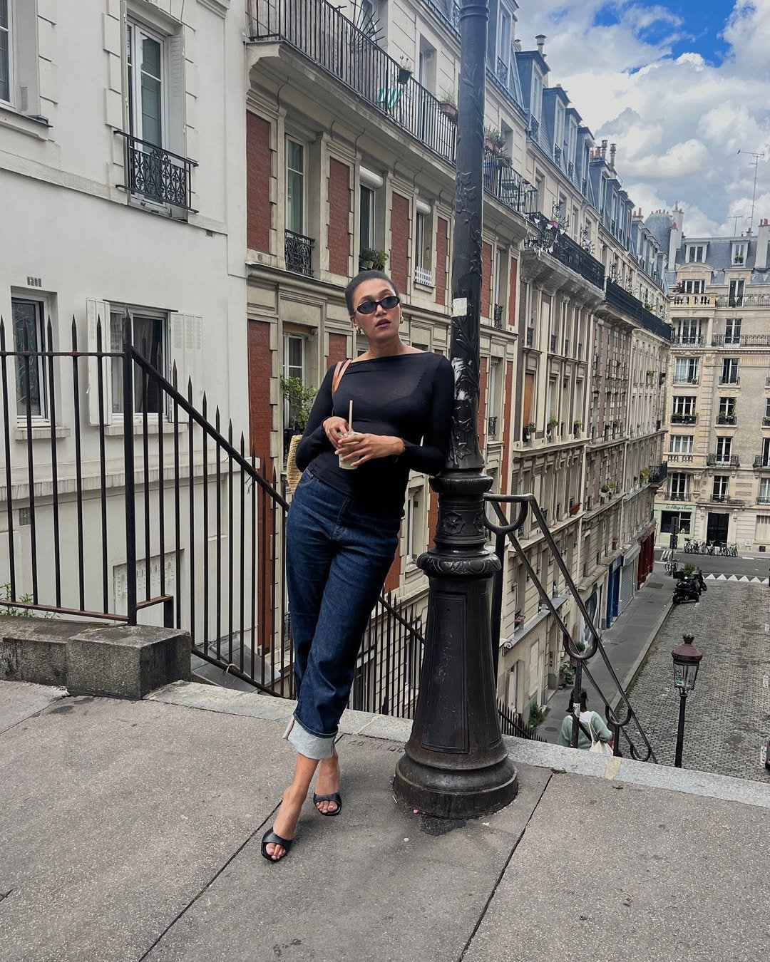Pessoa com blusa preta e calça jeans estilosa de 2025, apoiada em um poste, em cenário urbano europeu.
