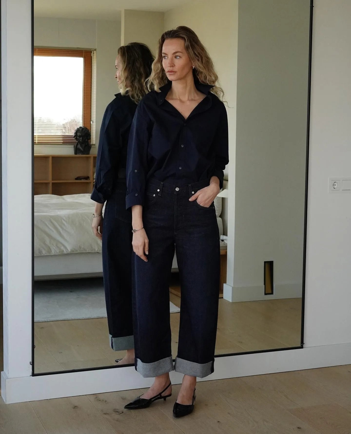 Mulher em frente ao espelho, vestindo camisa e calça jeans da moda de 2025, com barra dobrada e sapatos pretos.