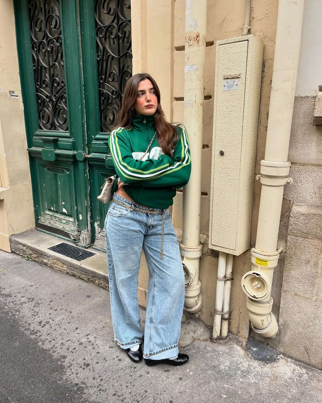 Pessoa vestindo calça jeans larga e jaqueta verde, encostada em parede com porta verde ao fundo.