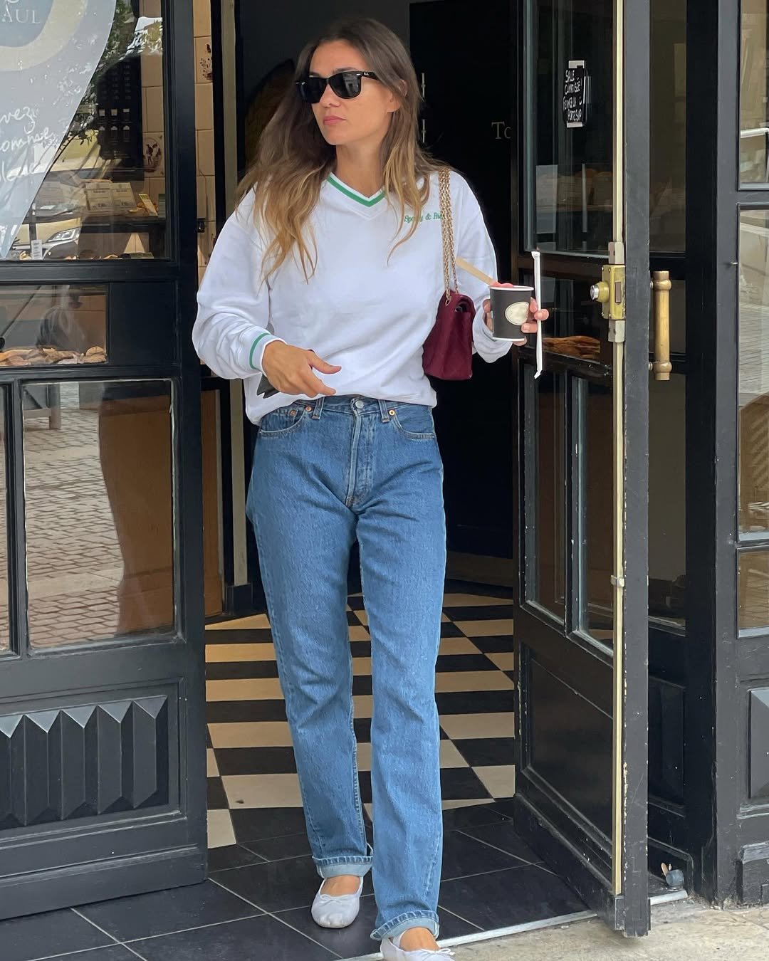 Pessoa saindo de loja usando blusa branca e calça jeans da moda de 2025, segurando café e bolsa vermelha.