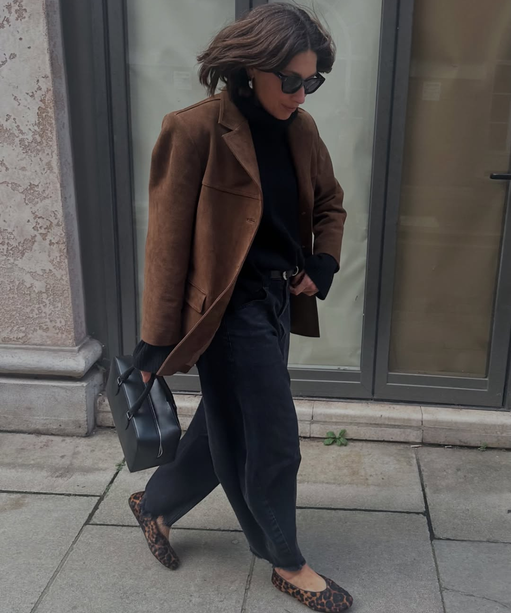 Mulher usa jaqueta marrom, suéter preto e calça jeans preta, segurando bolsa preta, sapatos com estampa animal.