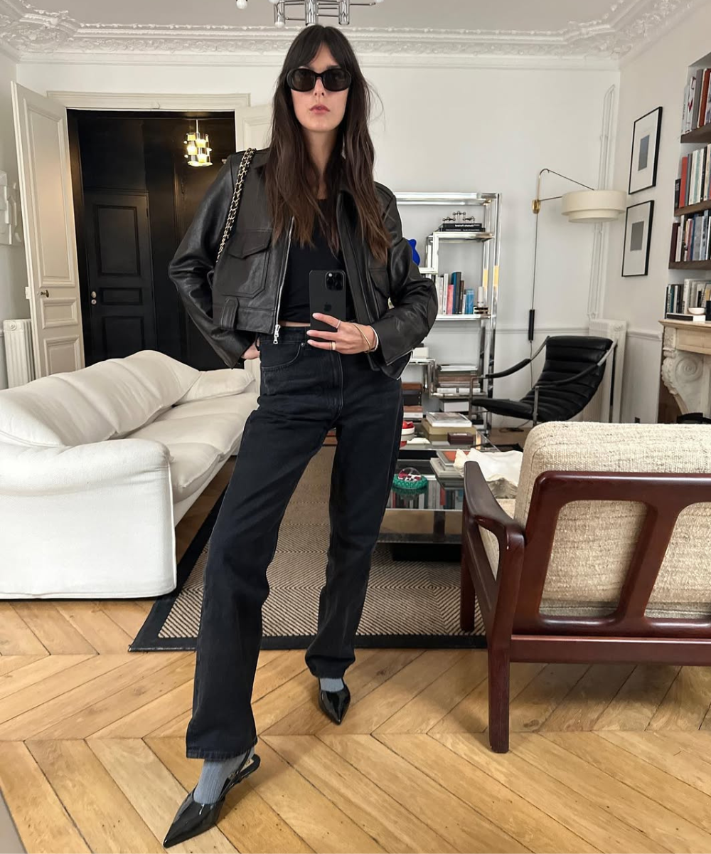 Mulher com jaqueta preta e calça jeans preta em sala moderna, segura celular e usa óculos escuros.