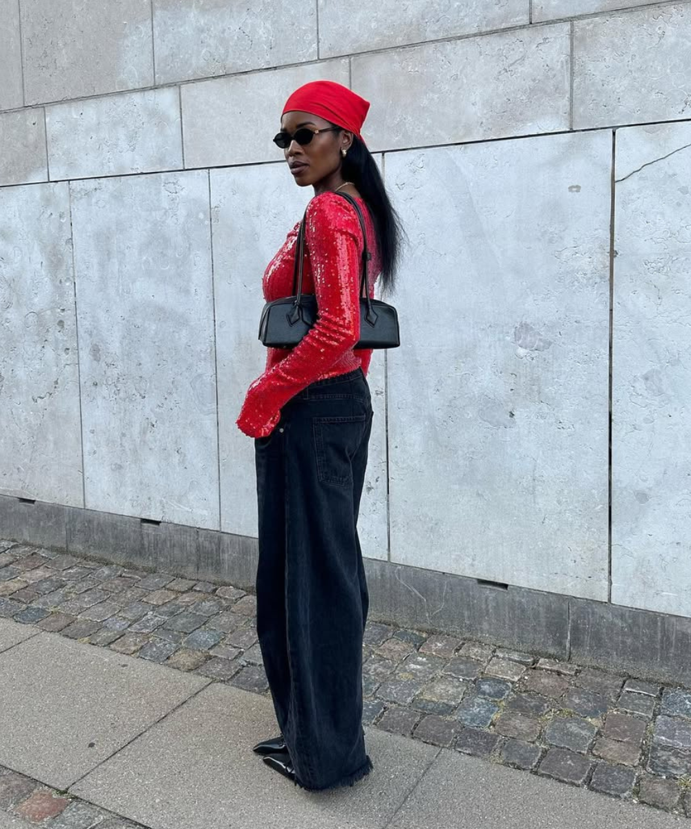 Pessoa de lenço vermelho e blusa brilhante, usando calça jeans preta, bolsa preta e óculos escuros.