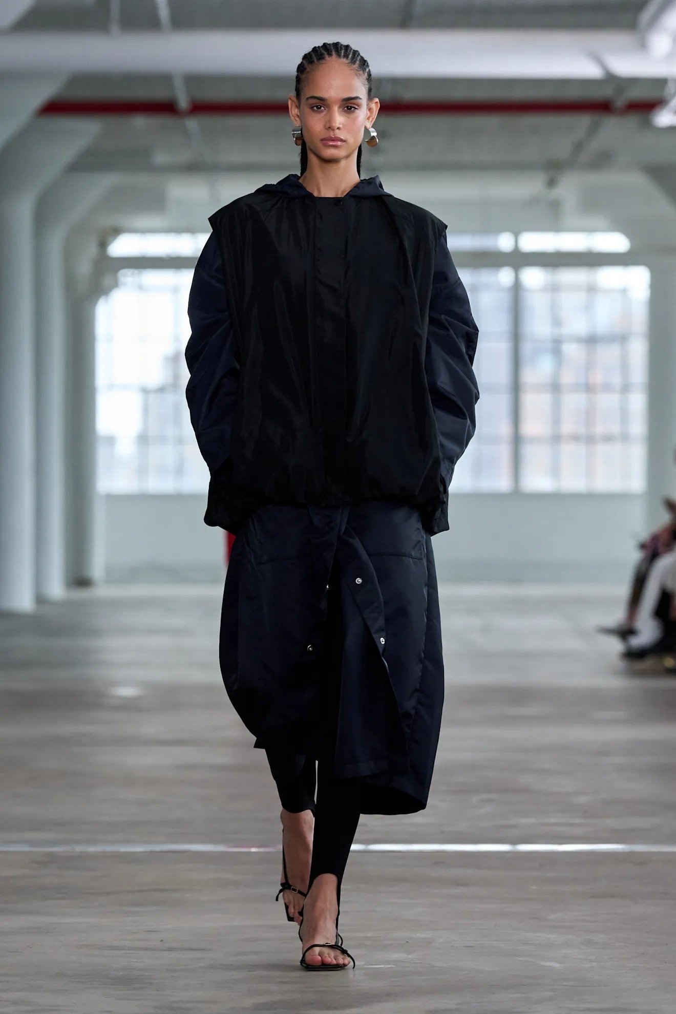 Modelo em desfile usando casaco preto longo, sandálias abertas e legging fusô preta. Fundo com janelas grandes.