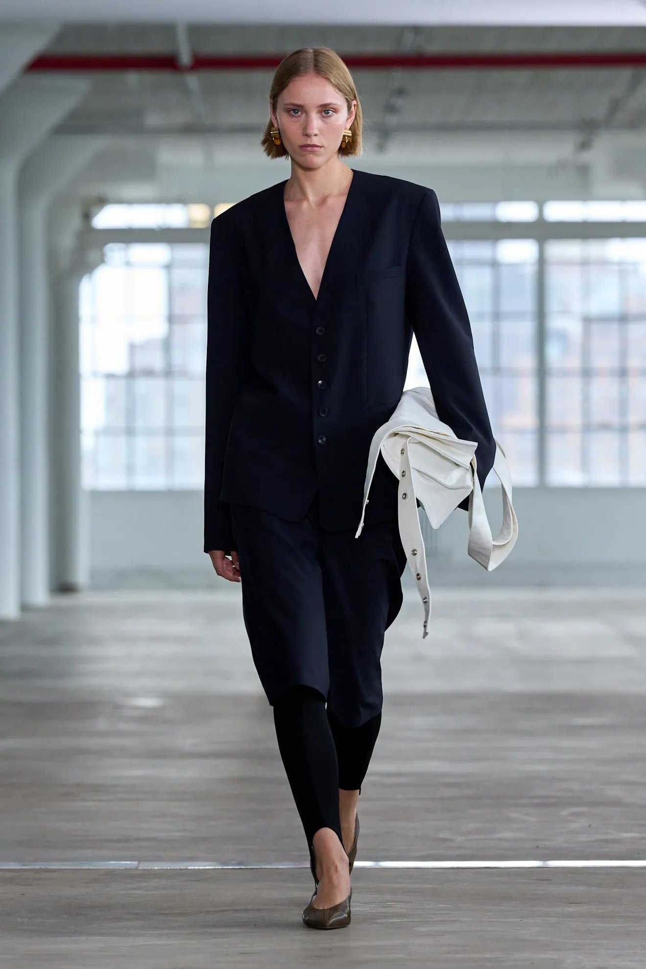 Modelo com blazer preto e legging fusô preta, segurando uma bolsa branca, em passarela minimalista.