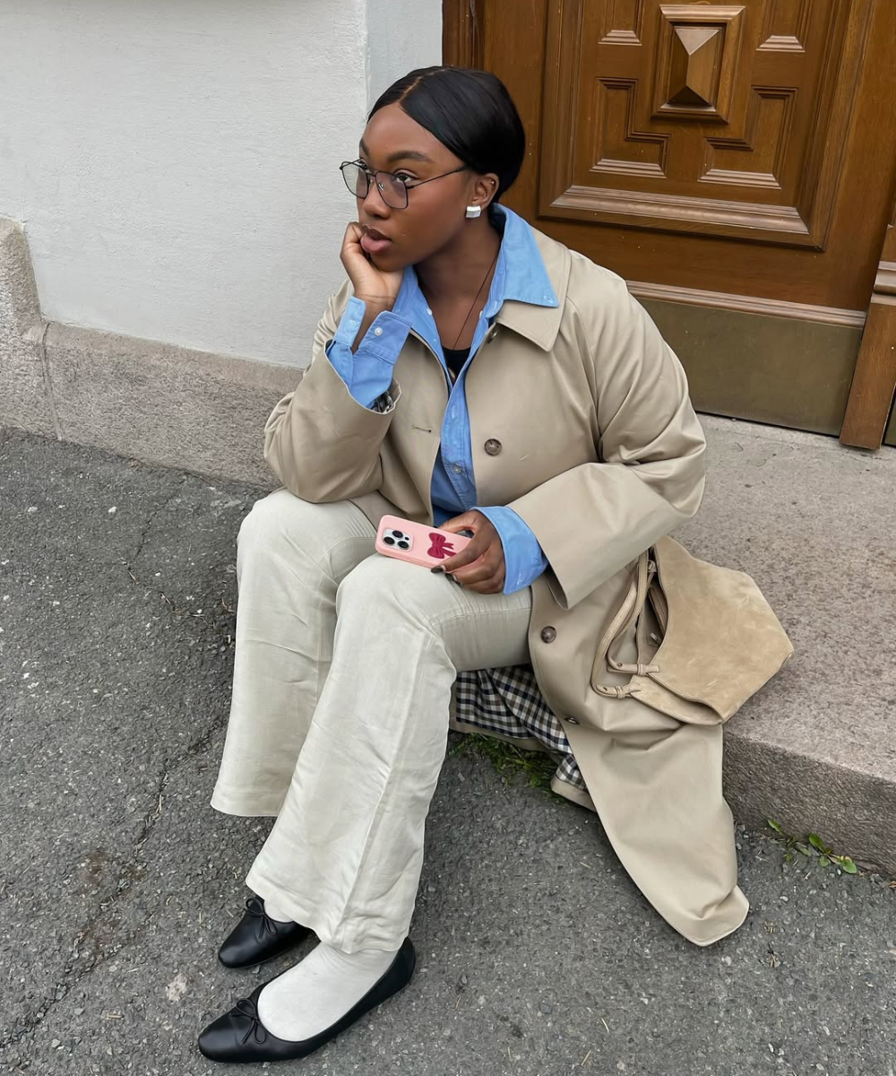 Pessoa sentada com casaco bege sobre camisa azul elegante. Ela segura um celular rosa, usando óculos e sapatilhas pretas.