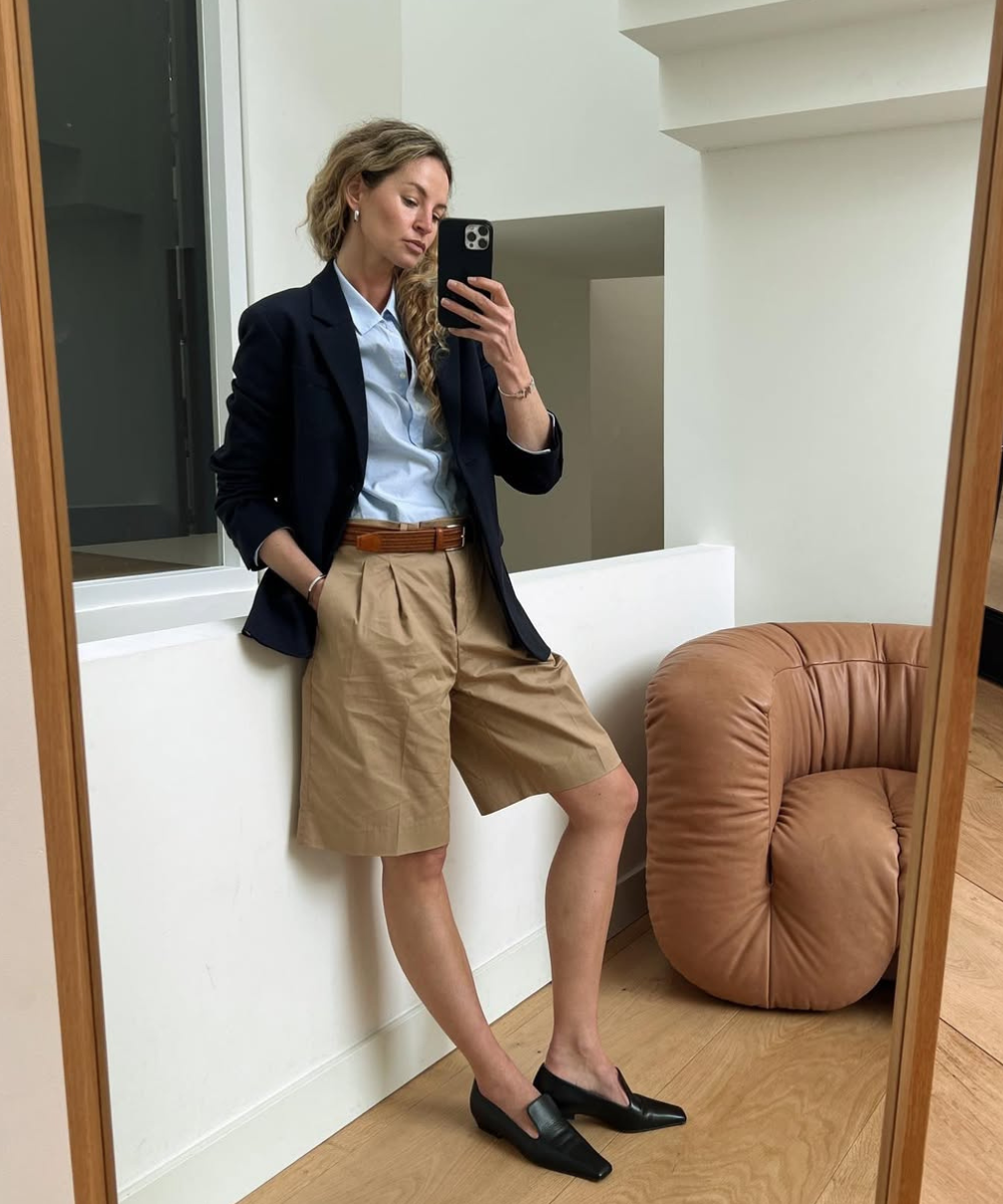 Pessoa com blazer sobre camisa elegante em um ambiente moderno, usando bermuda cáqui e mocassins pretos.
