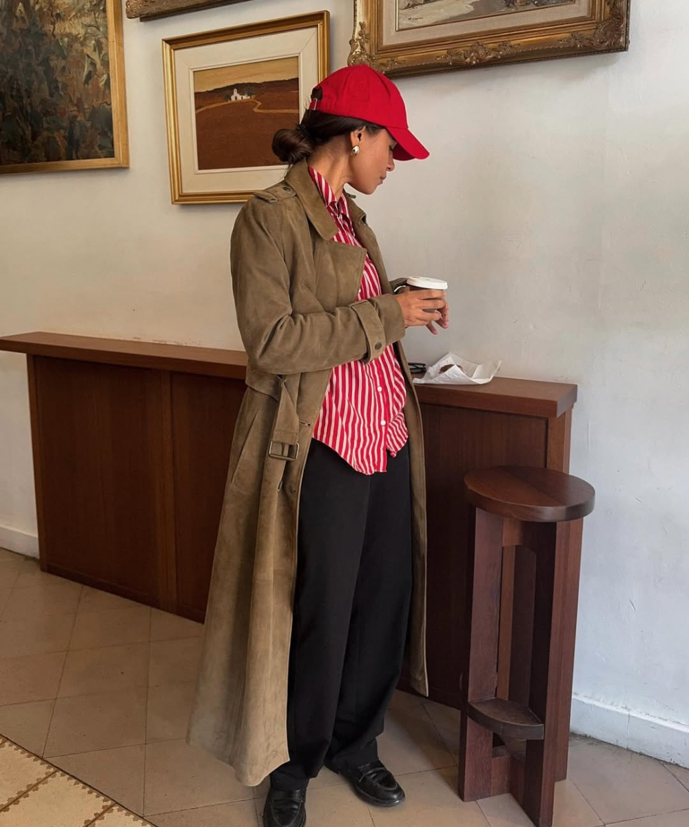 Mulher com camisa elegante listrada e casaco longo, usando boné vermelho, segura café em ambiente com quadros.