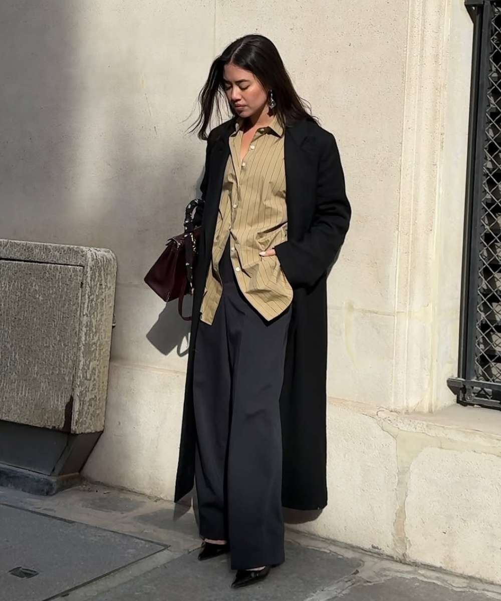 Mulher em pé usando camisa listrada elegante, calça preta e casaco longo, segurando bolsa marrom.