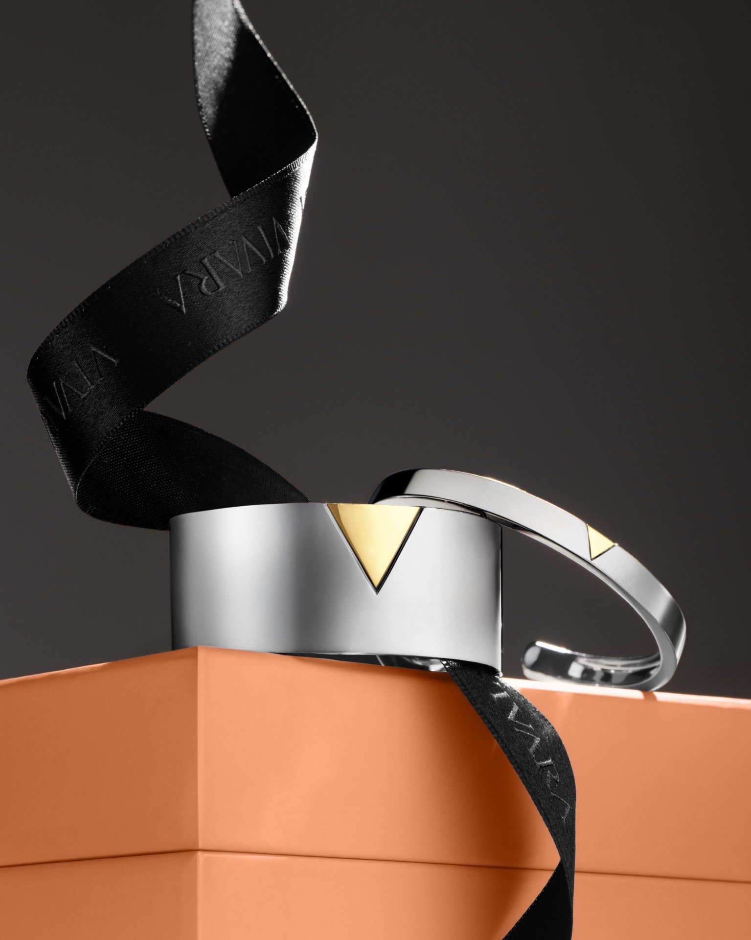 Braceletes metálicos prateados e dourados em caixa laranja com laço preto.