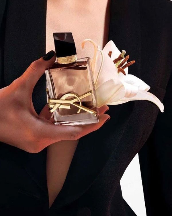 Frasco elegante de perfume Libre em formato retangular, segurado por uma mão feminina ao lado de uma flor branca.