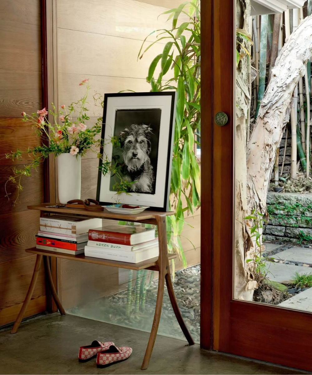 Mesa com livros, vaso de flores e foto de cachorro em uma casa com planta perto da porta de vidro.