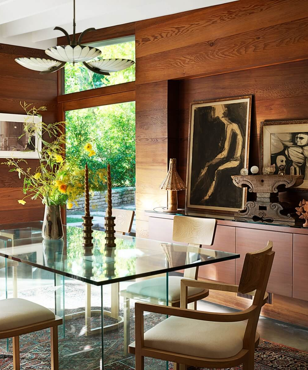 Casa da Dakota Johnson. Sala de jantar com mesa de vidro e cadeiras de madeira clara, decoração rústica e obras de arte nas paredes.