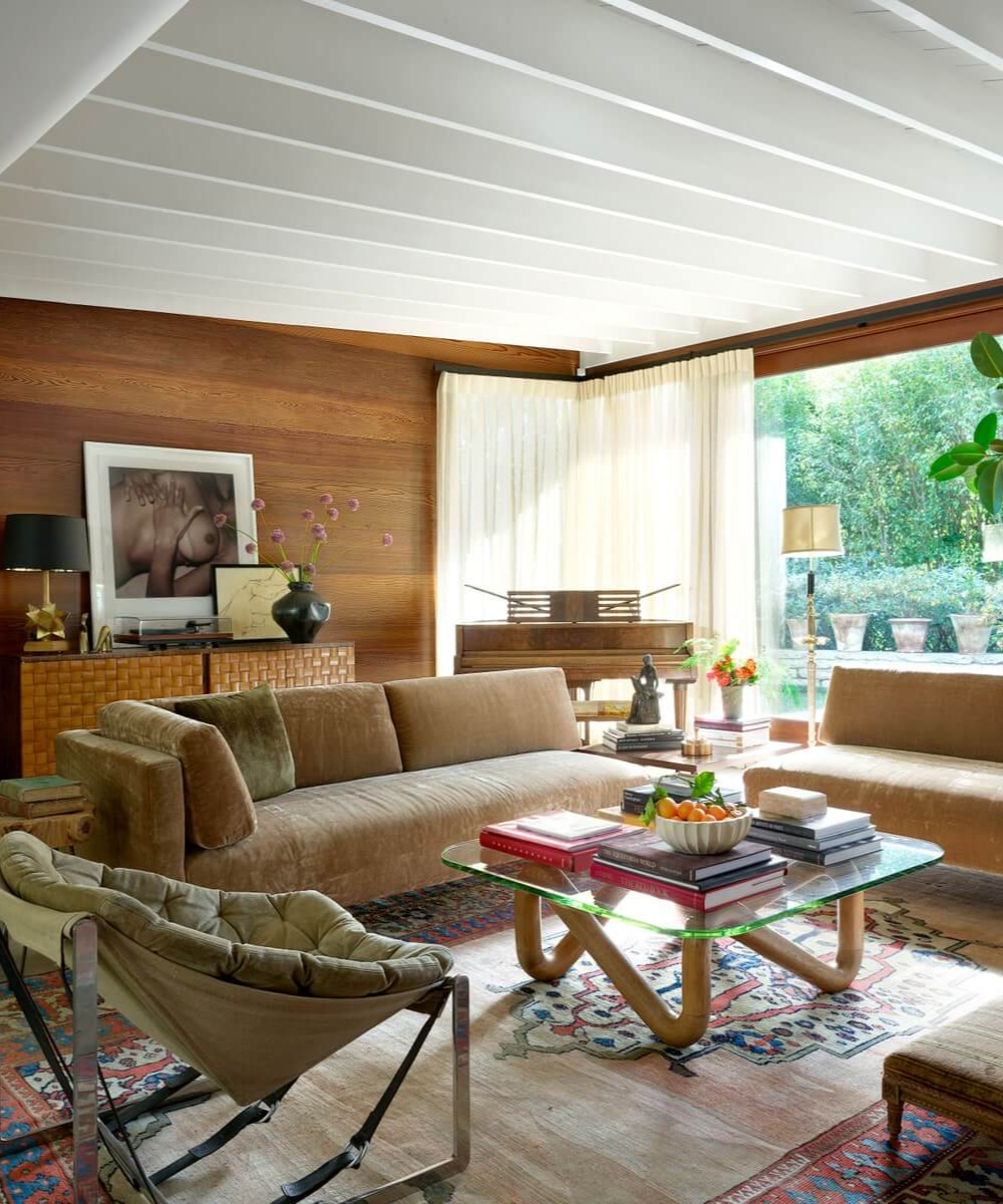 Sala elegante com sofás de veludo, mesa de vidro com livros, decoração estilosa e vista para jardim.