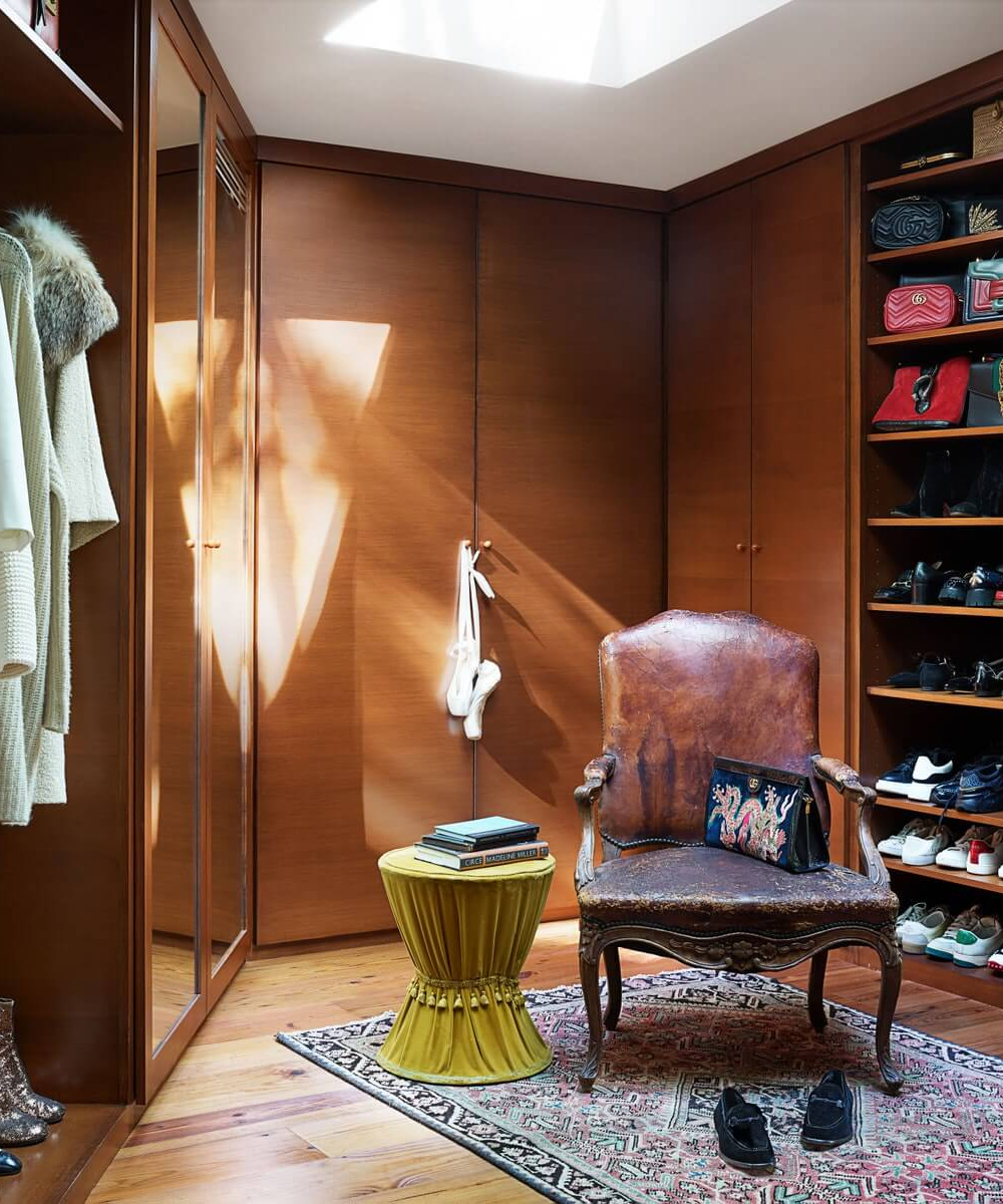 Closet da casa de Dakota Johnson com poltrona de couro, tapete estampado, mesinha com livros e sapatos organizados em estantes.