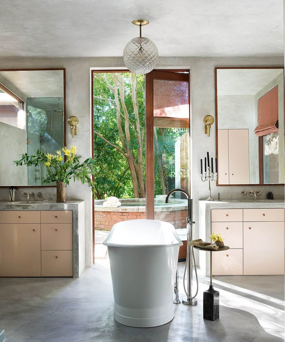 Sala de banho elegante com banheira central, armários claros e vista para jardim na casa da Dakota Johnson.