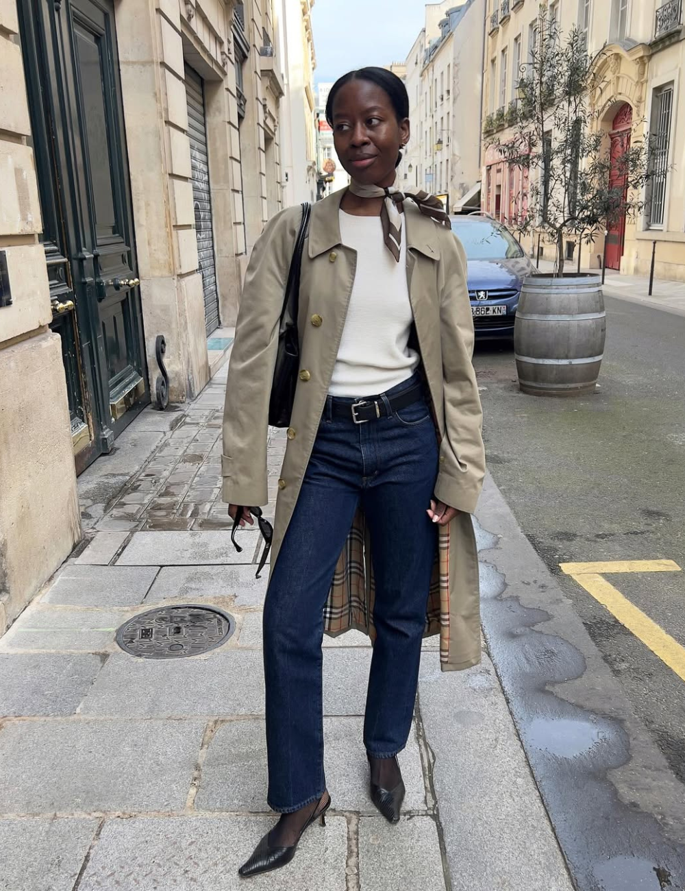 Pessoa em rua, veste um casaco clássico bege, jeans escuro e sapatos pretos, com uma bolsa e lenço no pescoço.