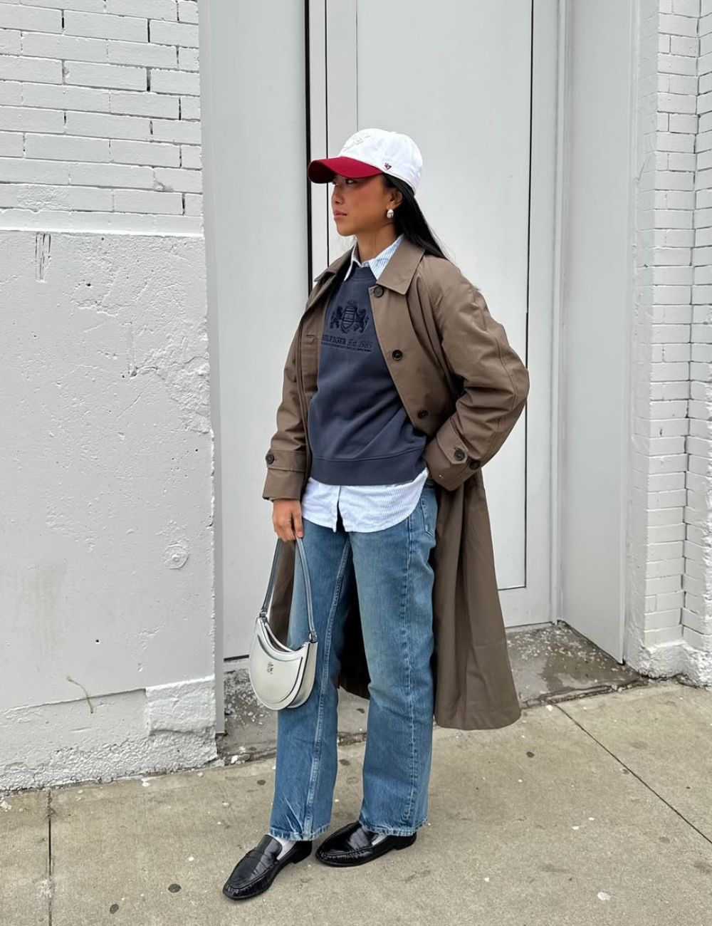 Pessoa usando casaco longo, jeans, suéter e boné branco com detalhe vermelho, segurando bolsa pequena e vestindo sapatos pretos.