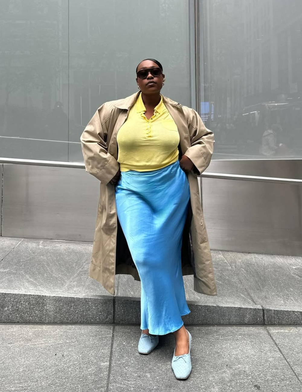 Pessoa usando um casaco clássico bege, blusa amarela e saia azul brilhante, posando ao ar livre.