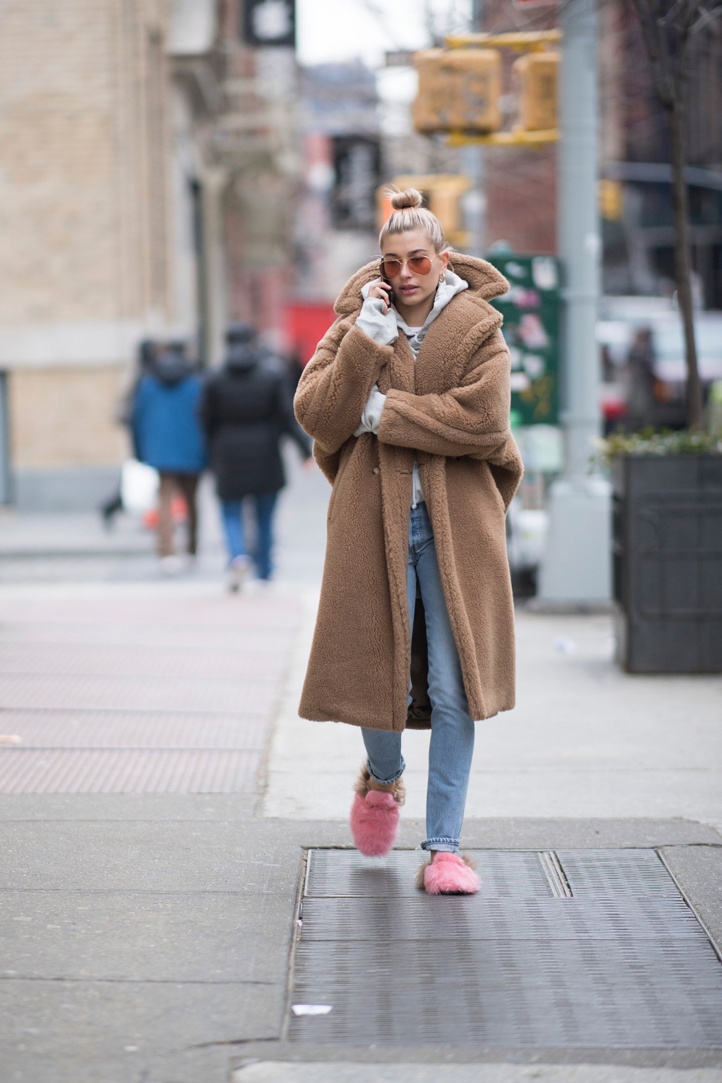 Pessoa usando um casaco teddy marrom, calça jeans e chinelos rosa em uma calçada urbana.