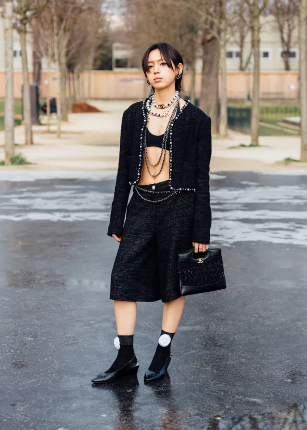 Pessoa em look preto elegante com joias chamativas, segurando uma bolsa. Árvores ao fundo em ambiente urbano.