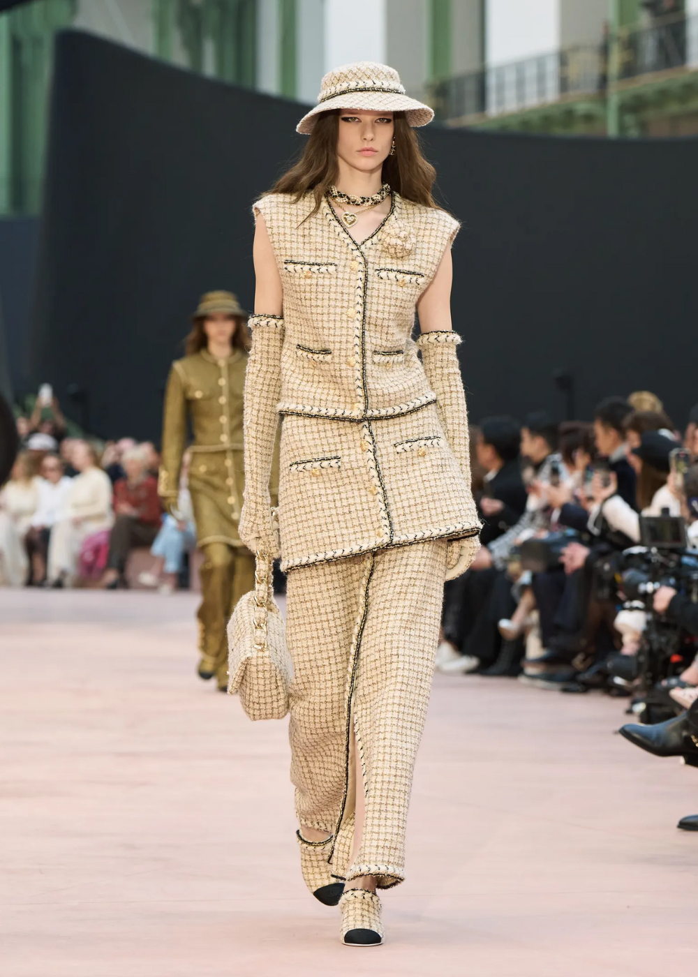 Modelo desfila em uma passarela, usando traje de tweed claro com chapéu e luvas combinando. Público ao fundo. Foto para ilustrar o nevold.