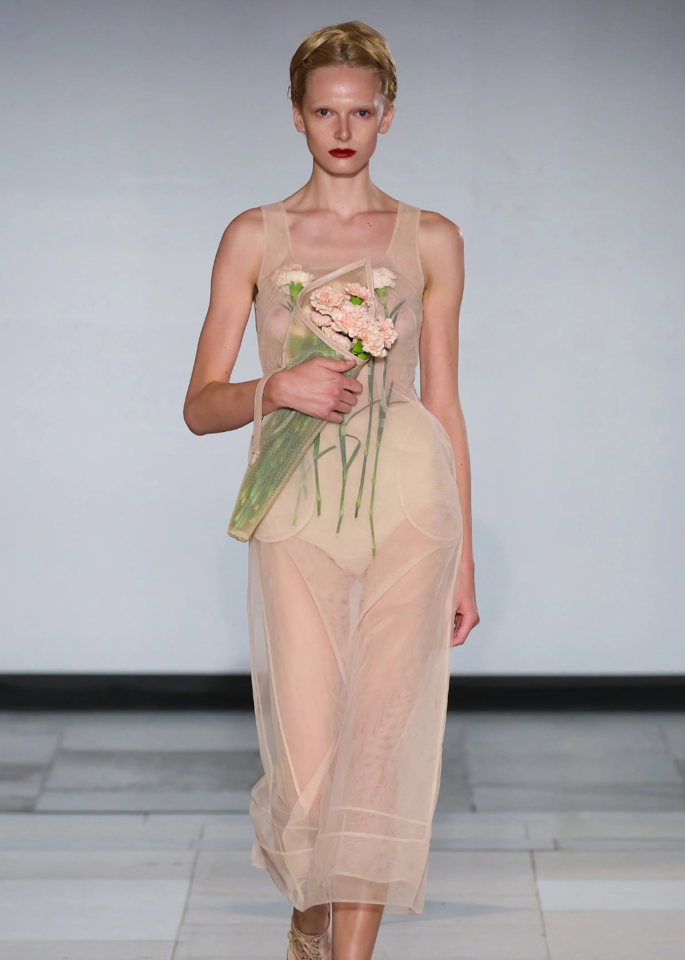 Desfile de moda com modelo usando vestido translúcido, segurando flores.