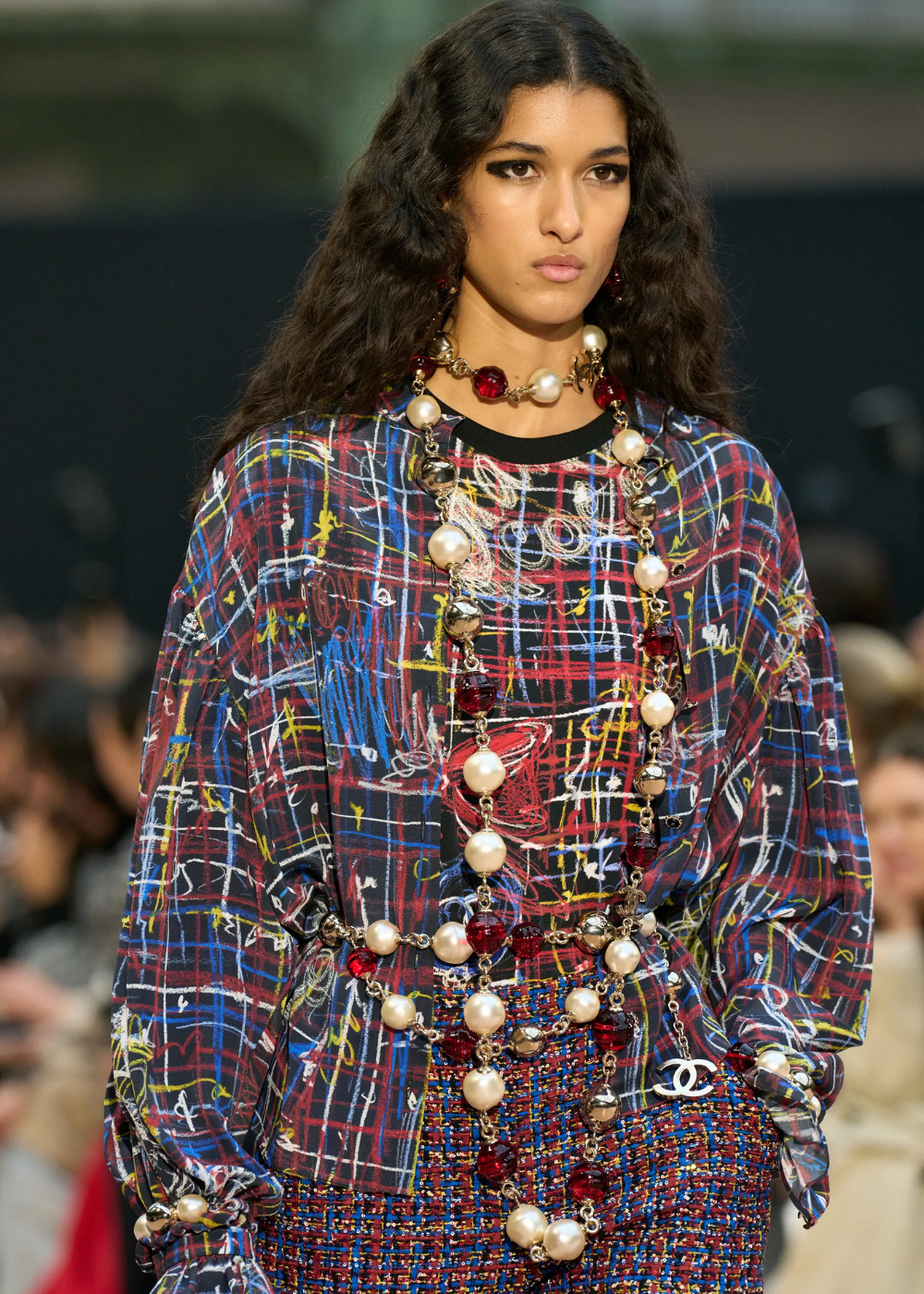 Modelo veste blusa colorida com colar de pérolas e pedras, em desfile de moda.
