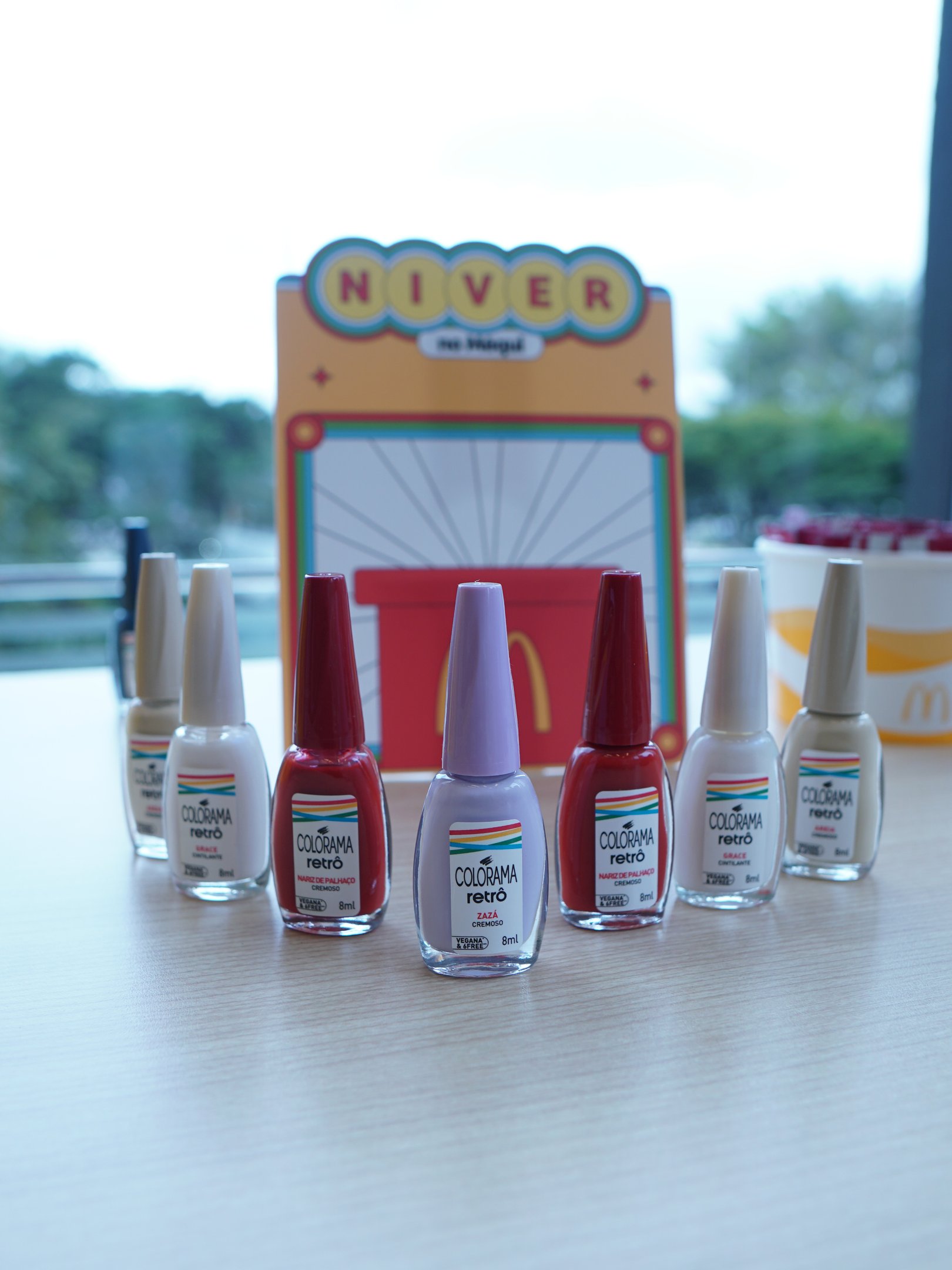 Esmaltes da coleção retrô Colorama em diferentes tons, dispostos em linha sobre uma mesa. Ao fundo, decoração comemorativa.