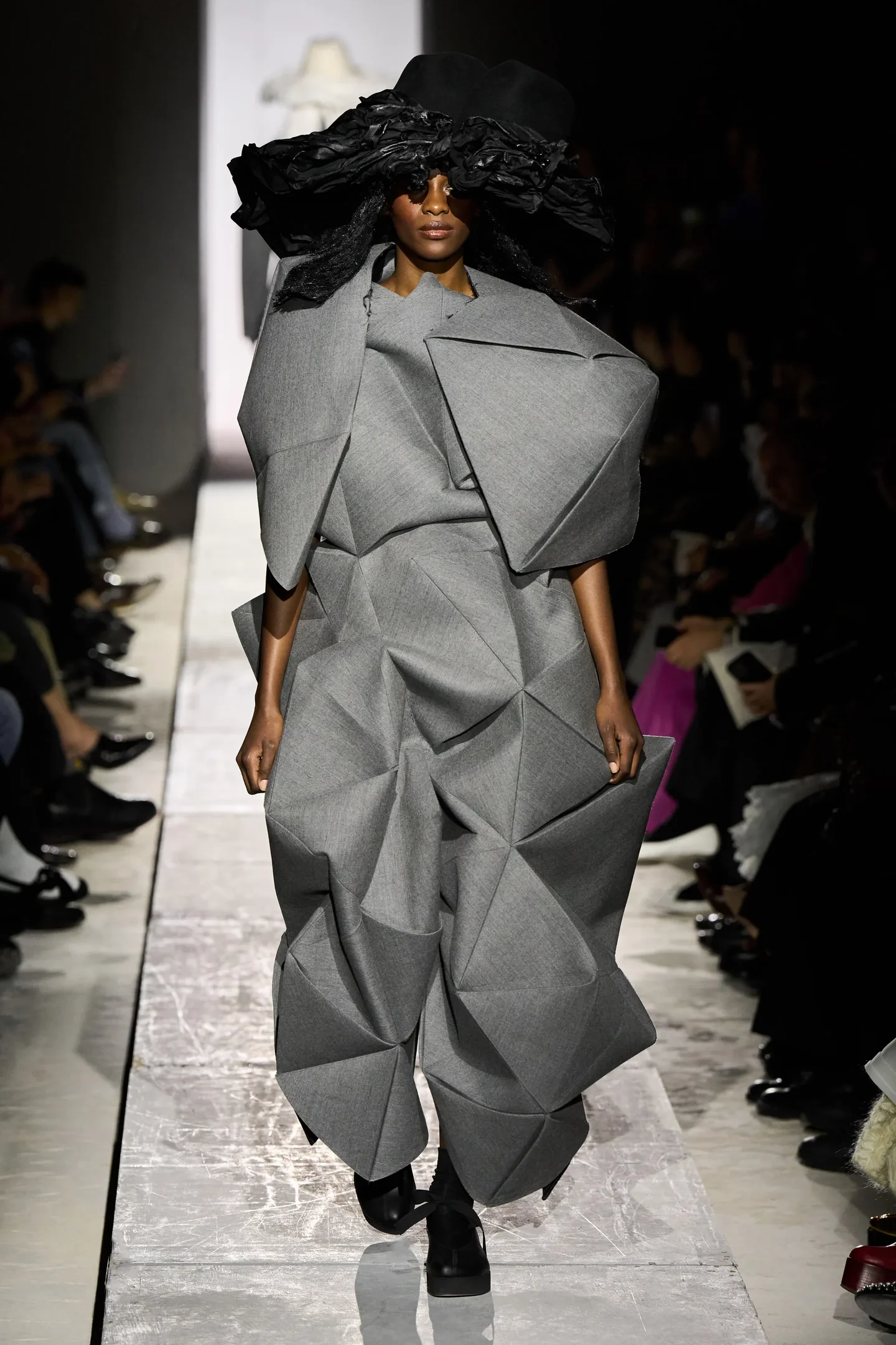 Modelo em passarela, usando traje cinza geométrico de Rei Kawakubo, com chapéu preto volumoso.