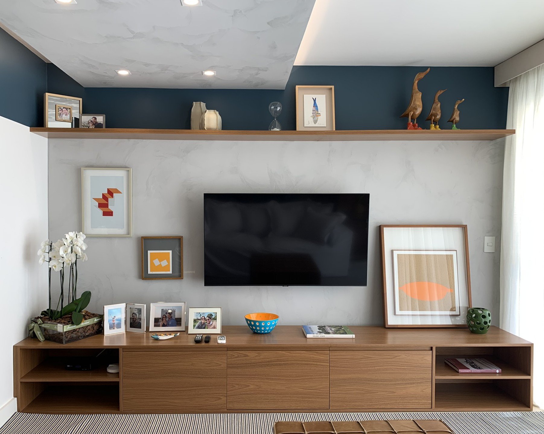 Como decorar sala pequena com prateleira, TV montada na parede, quadros, orquídeas e objetos decorativos.