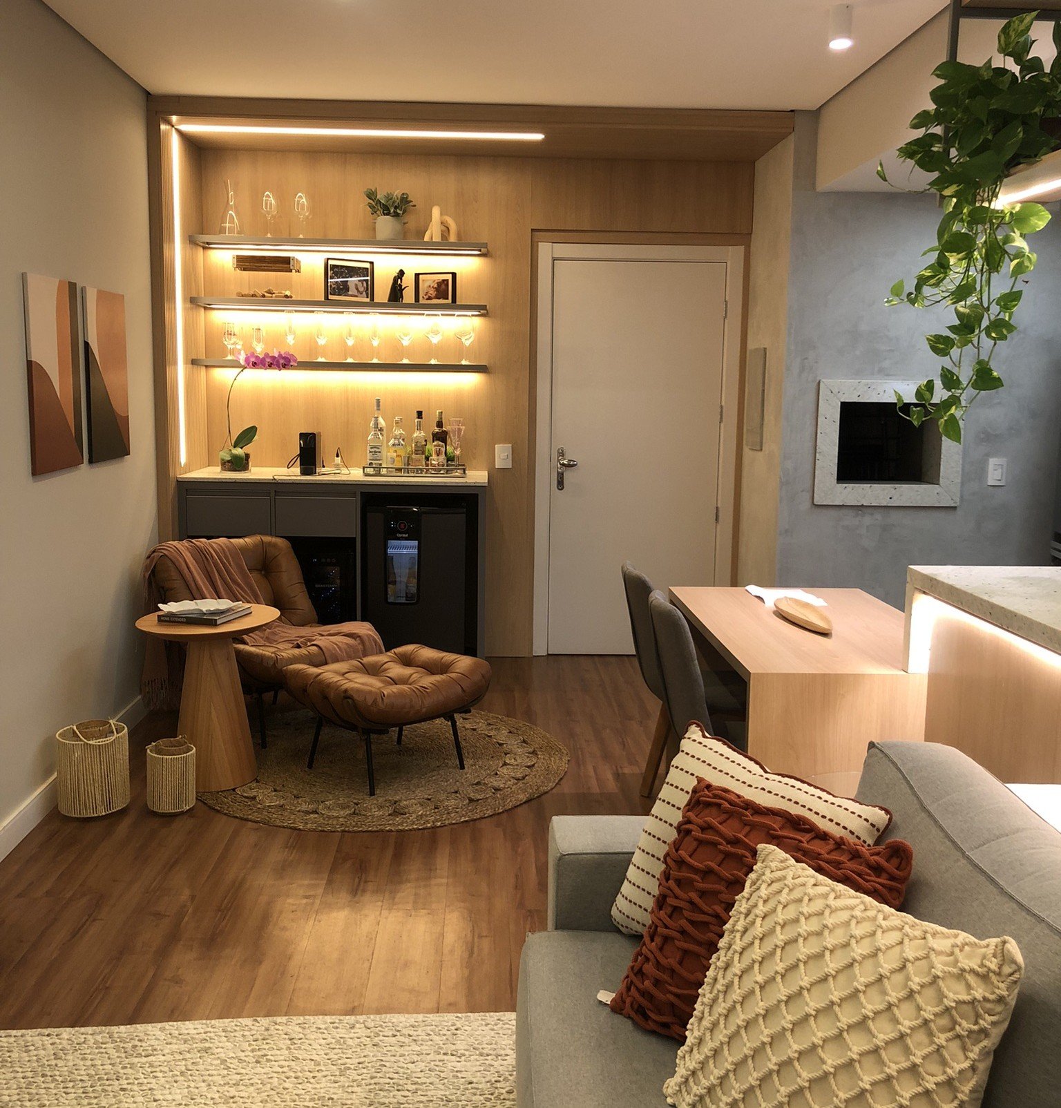 Sala pequena bem decorada com estante iluminada, poltrona confortável, plantas e mesa de jantar compacta.