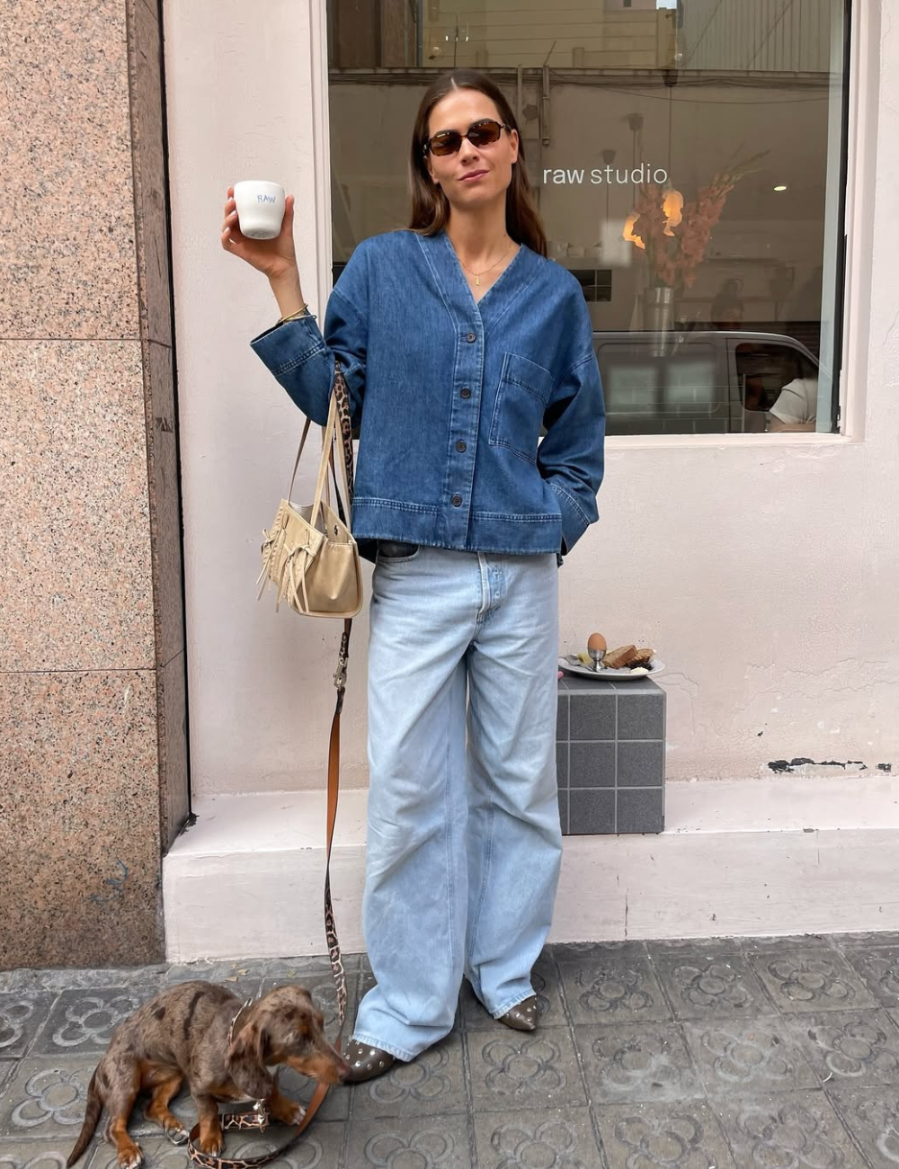 Pessoa com jaqueta jeans segura café, vestindo calça jeans baggy. Cão ao lado na calçada.