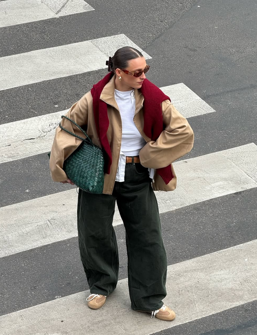Pessoa usando calça jeans baggy e casaco bege. Blusa branca e suéter vermelho nos ombros. Bolsa verde e óculos escuros completam o look.