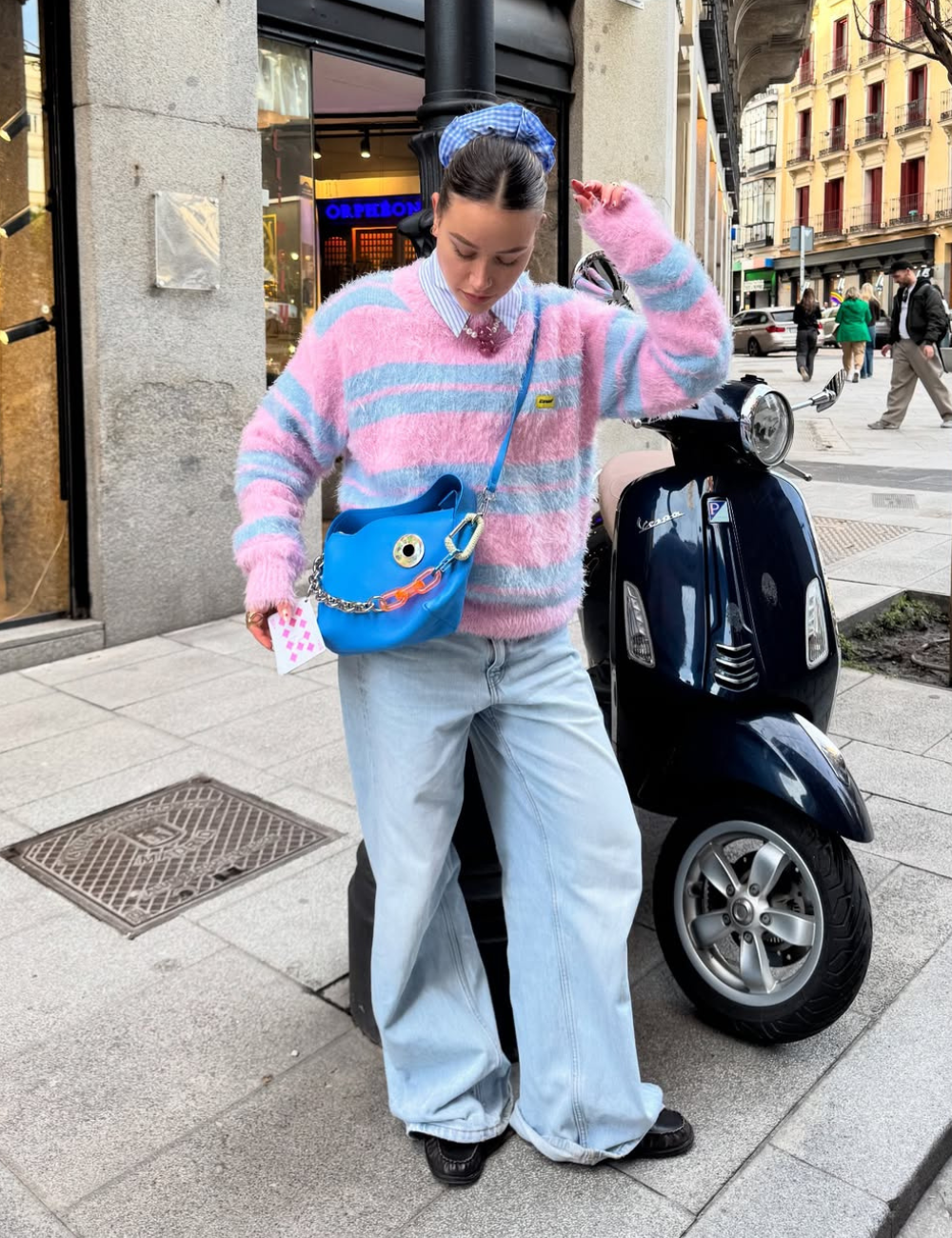 Pessoa vestindo suéter listrado rosa e azul, calça jeans baggy e segurando bolsa azul, próxima a uma moto.