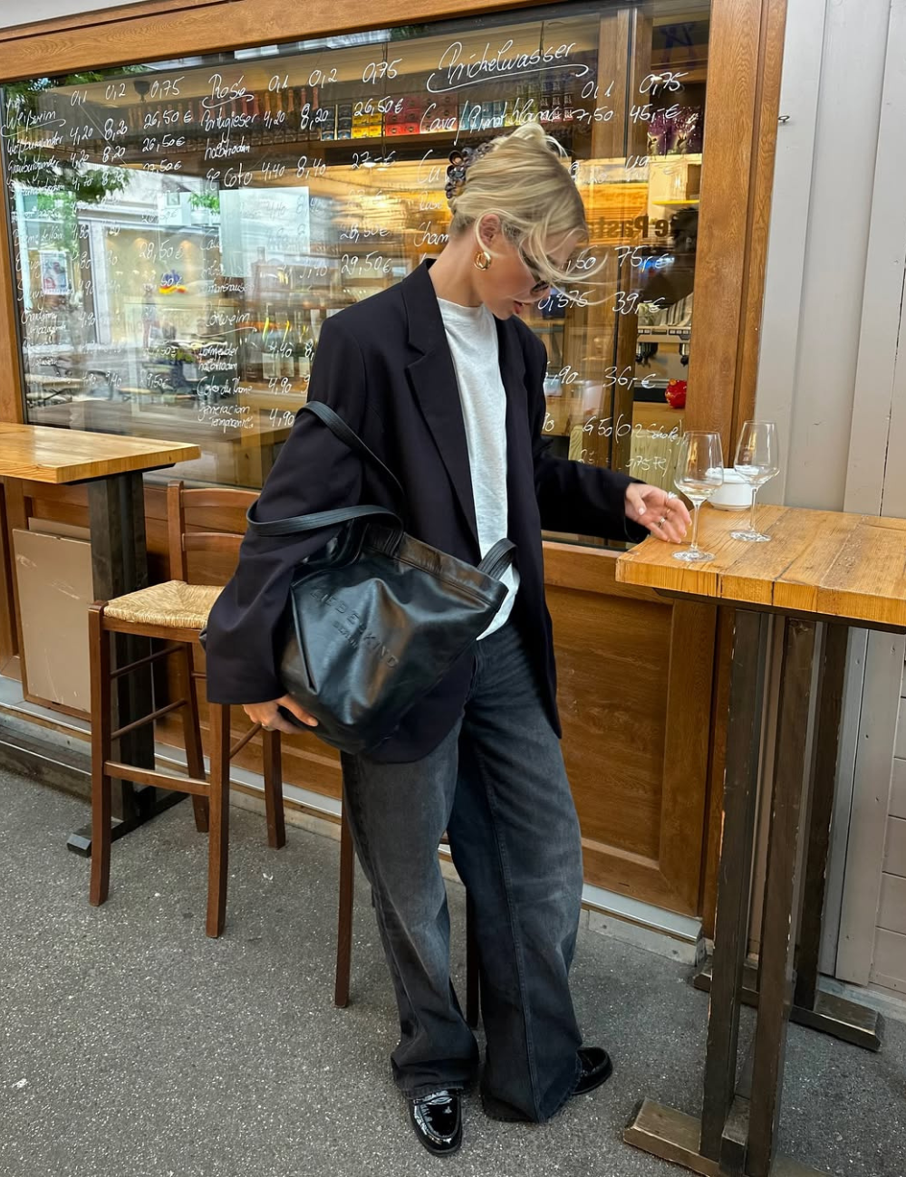 Pessoa vestindo blazer preto e calça jeans baggy, segura bolsa grande em frente a um bar com mesas altas.