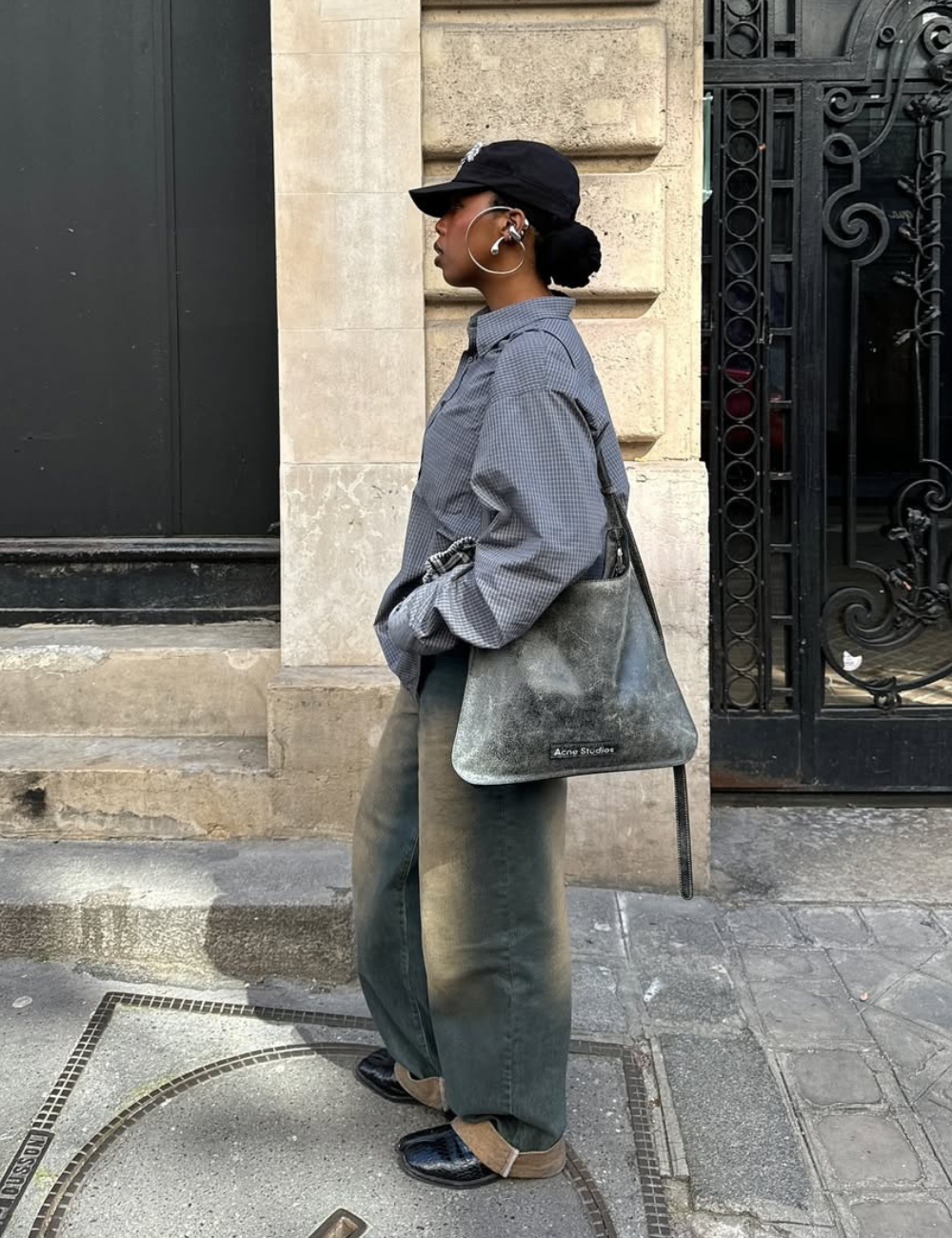 Pessoa usando calça jeans baggy, camisa larga, boné preto e bolsa cinza, caminhando por uma calçada urbana.