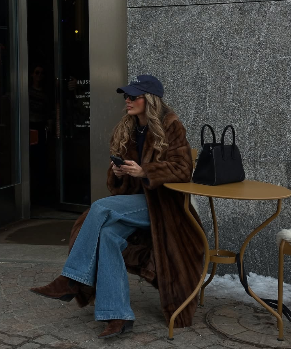 Pessoa sentada em uma mesa ao ar livre, usando casaco marrom comprido, calça jeans e chapéu azul.