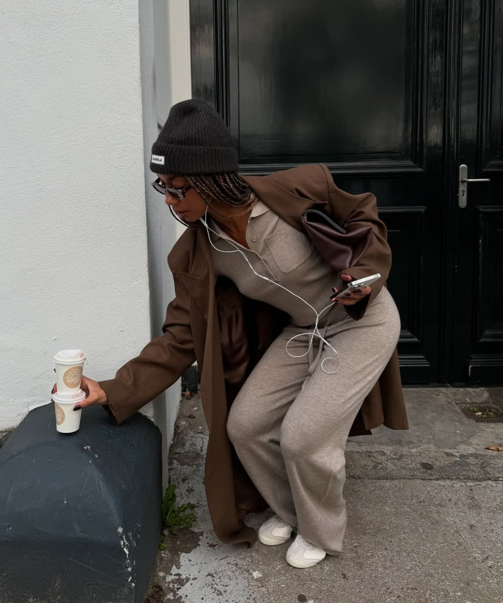 Pessoa usando casaco marrom e gorro escuro, segurando café e celular diante de uma porta preta. Mostra como usar marrom no inverno.