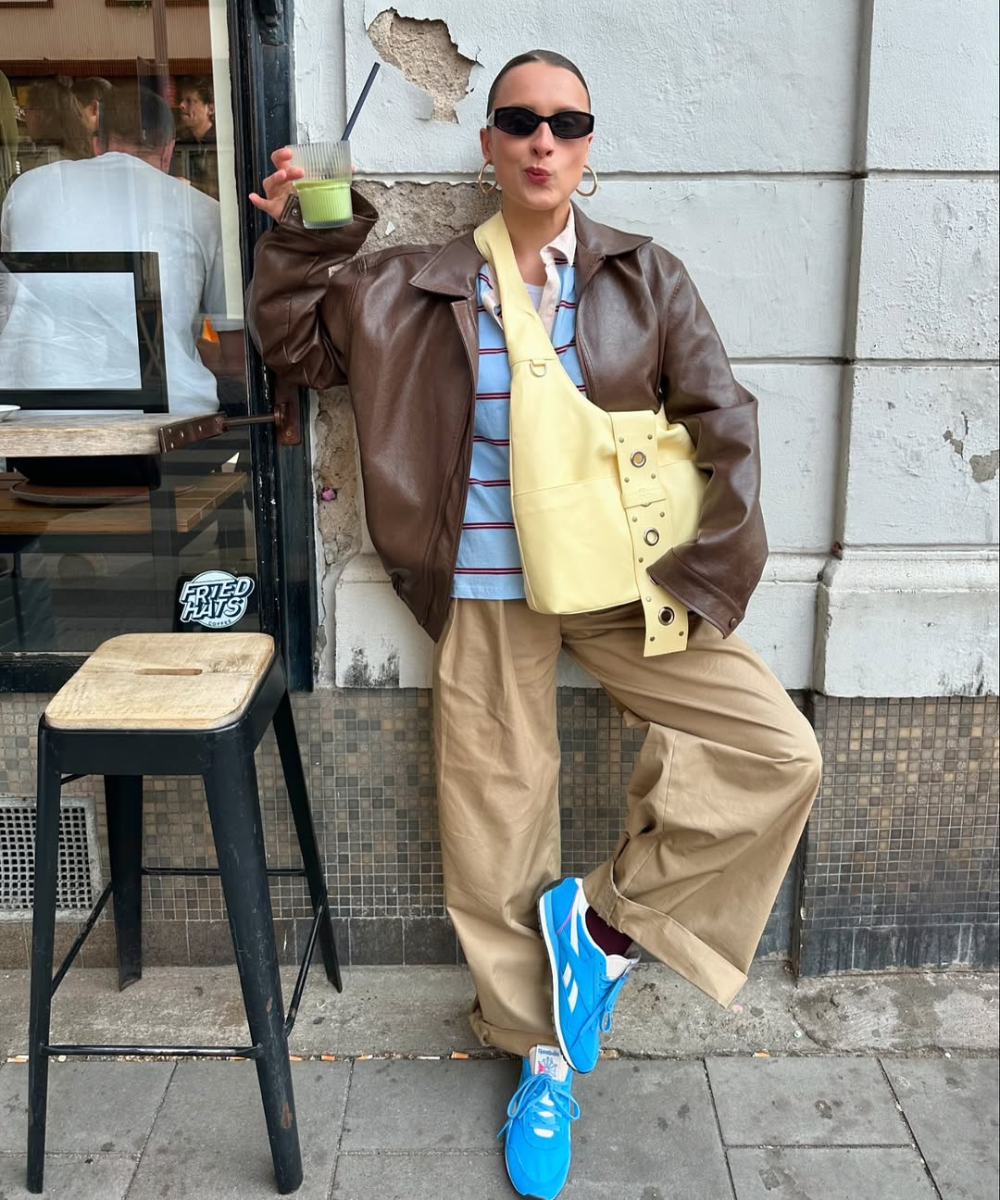 Pessoa usando casaco marrom, calça caqui, tênis azul e segurando uma bebida verde. Bolsa amarela no ombro.