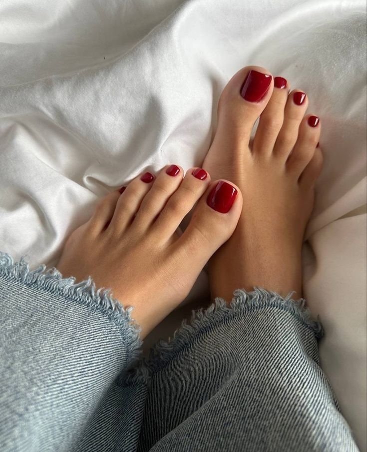 Unhas dos pés esmaltadas em vermelho escuro, em uma pessoa usando jeans desfiado, sobre lençóis.