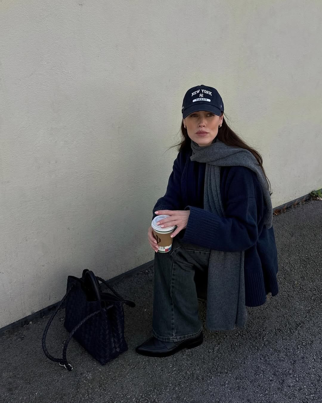 Pessoa usando boné e cachecol escuros, segurando um copo de café, com bolsa ao lado. Cores elegantes compõem o visual.