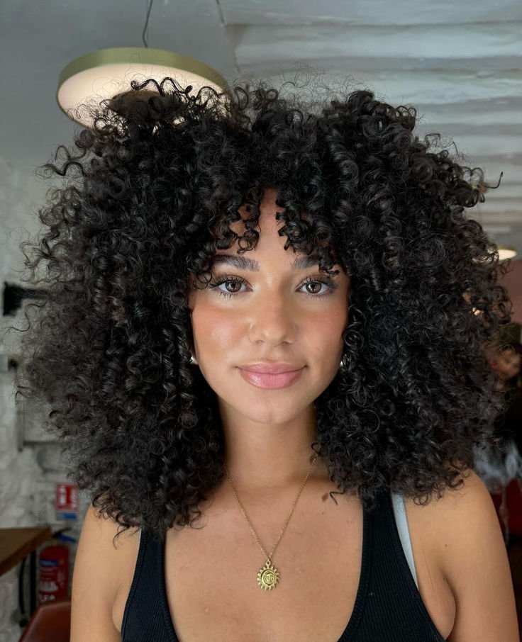 Jovem com cabelo cacheado volumoso, comprimento até os ombros, usa colar dourado, em ambiente interno iluminado.