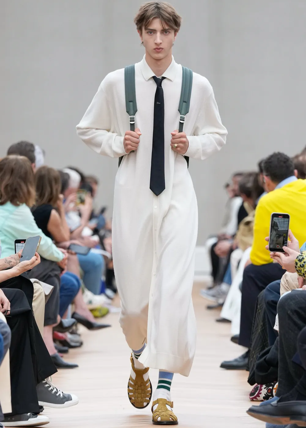 Modelo em passarela usa traje longo com sandália, mochila e gravata. Público ao redor fotografa.