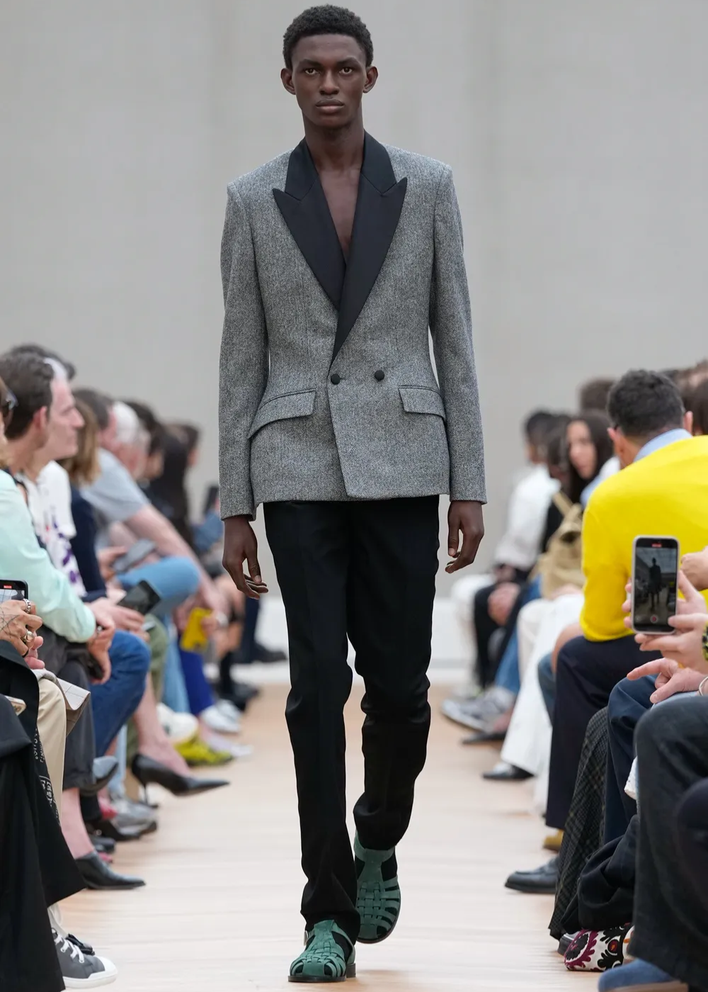 Modelo em passarela veste terno cinza e calça preta, usando sandálias verdes em desfile.